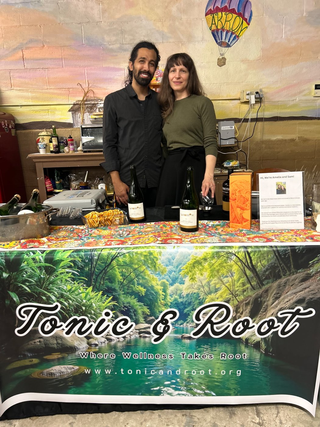The Tonic & Root team at a community pop-up, with bottled tonics on the table and the Tonic & Root banner behind them